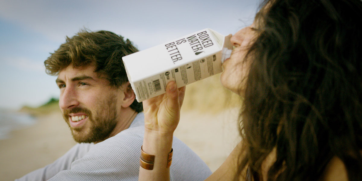photo showing a man and a woman sitting on the beach with woman drinking from a carton